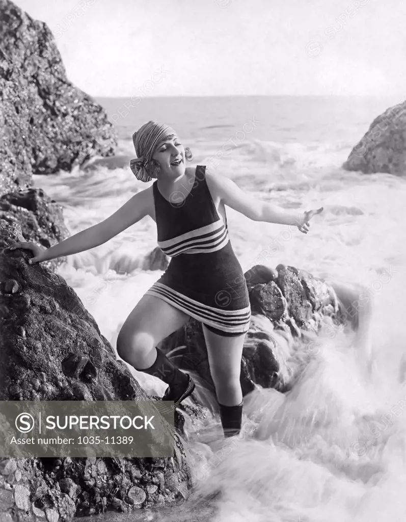 Los Angeles, California:  c. 1922. A flapper in a bathing suit, scarf, and footwear plays in the surf on a rock at the beach.©Underwood Archives