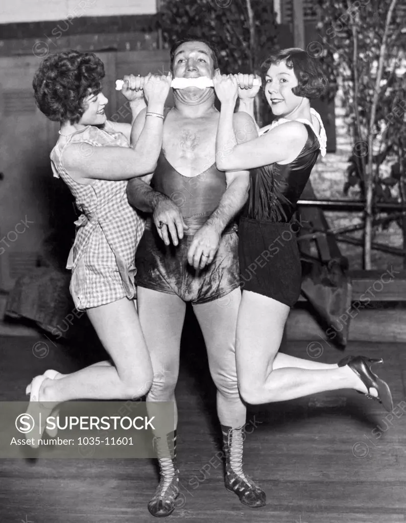 New York, New York:  December 29, 1927. Edward Reece, famous strongman, shows his power by holding up two women with his teeth.