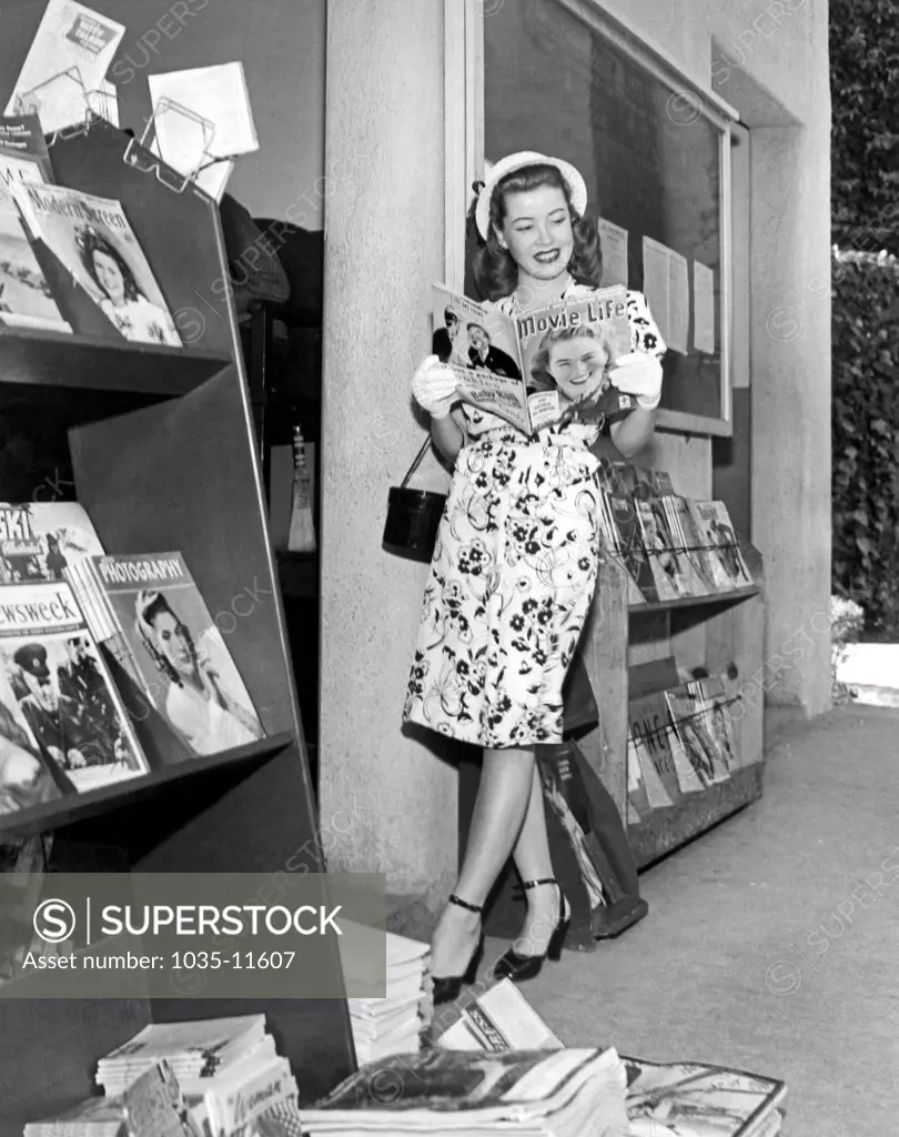 Hollywood, California:   April, 1945. A stylish young woman wearing a white hat and  gloves peruses the latest edition of 'Movie Life' at a magazine stand.