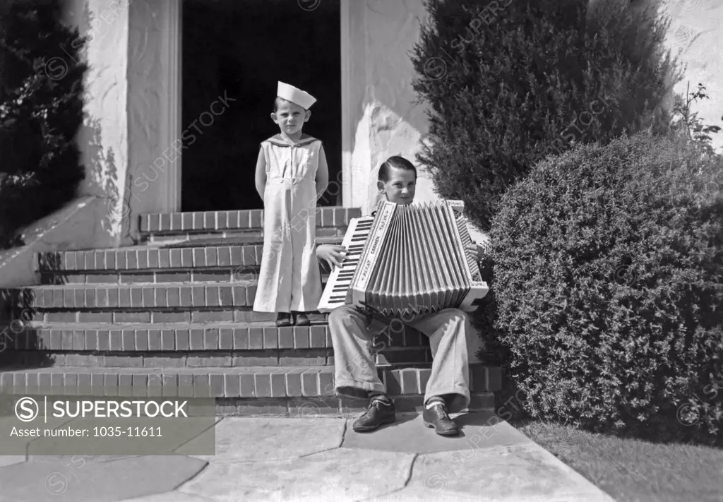 United States:  c. 1910  A little boy in a sailor's outfit stands on his front steps next to an older boy playing a large accordian.