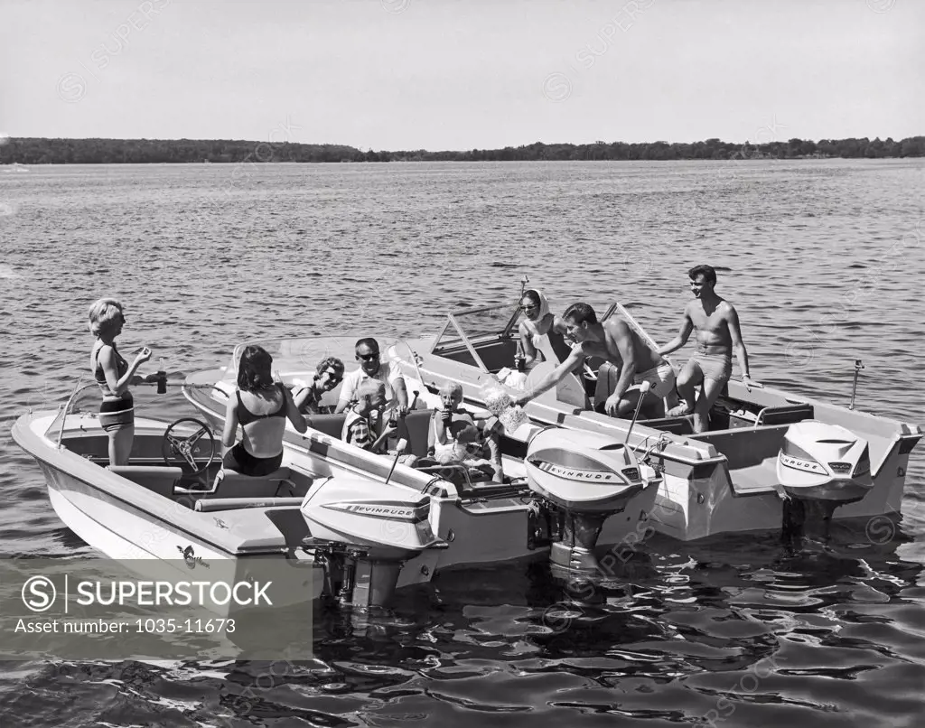 United States:  c. 1960. Three sets of boaters with Evinrude motors gather together on the lake to share food and drink.