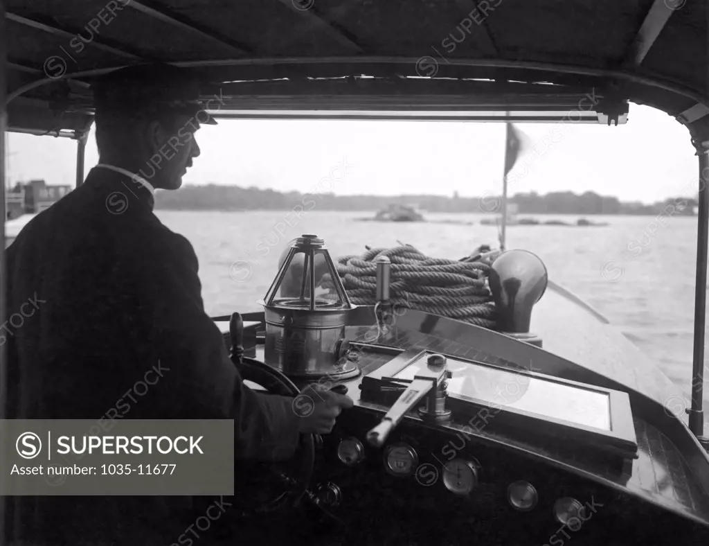 United States:   c. 1925. A man in a covered cockpit guides his large power boat up a waterway