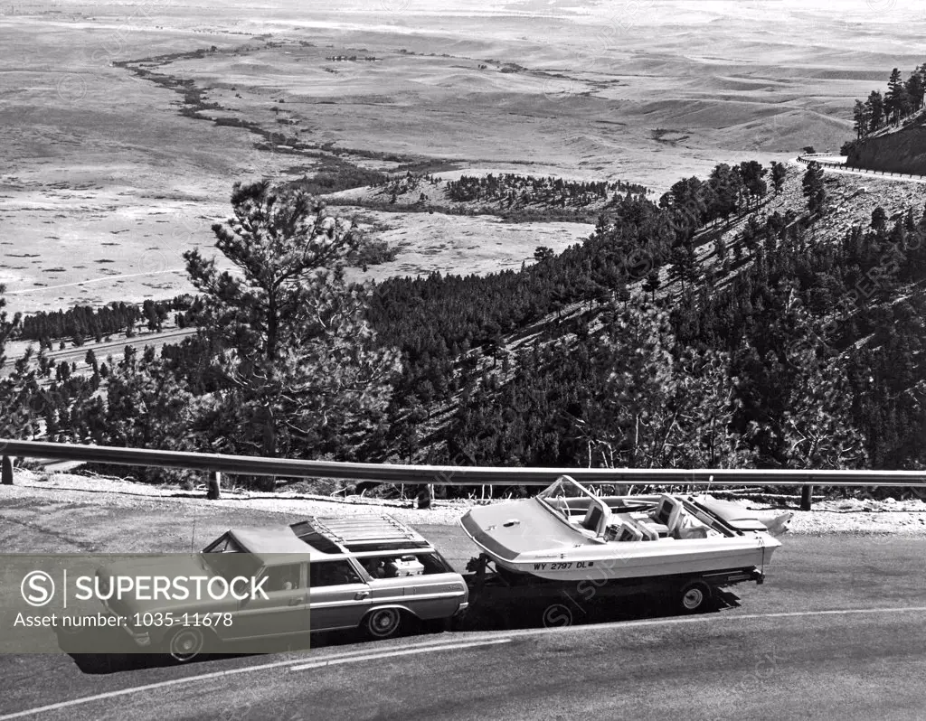 United States:  c. 1960. A station wagon towing a power boat heads up into the mountains from the valley below in the background.
