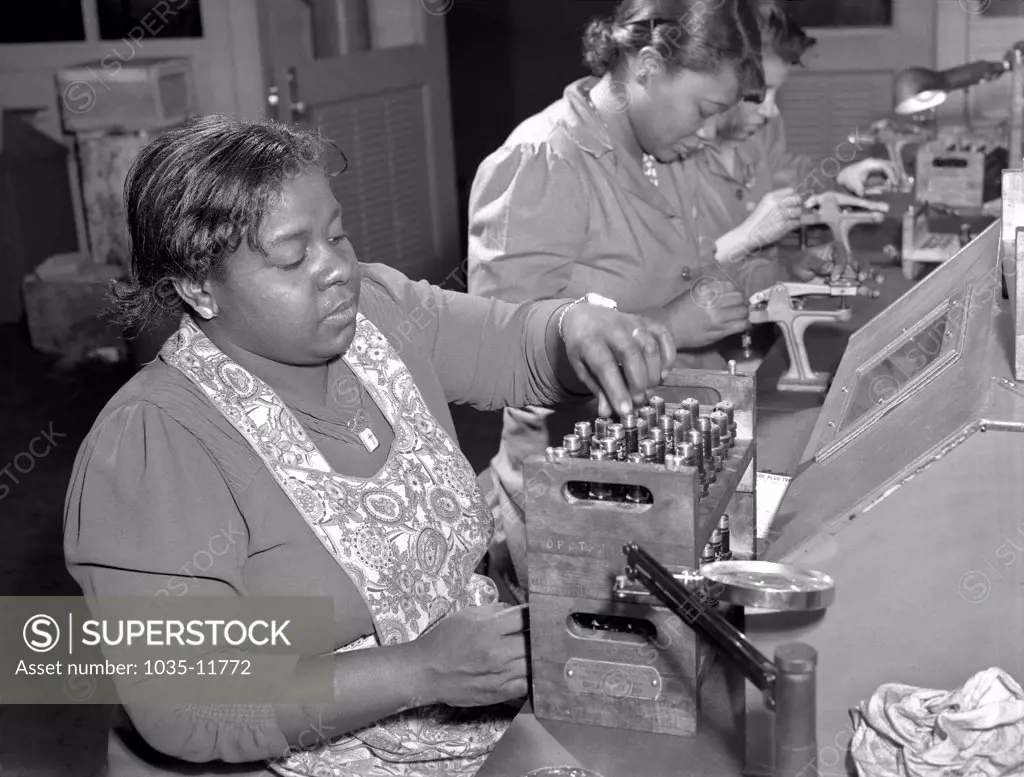 Melrose Park, Illinois:  February 1942. Negro women with no previous industrial experience are reconditioning used spark plugs in a converted Buick plant to produce airplane engines. Despite their lack of technical knowlege, these women have become expert operators of the small testing machines.