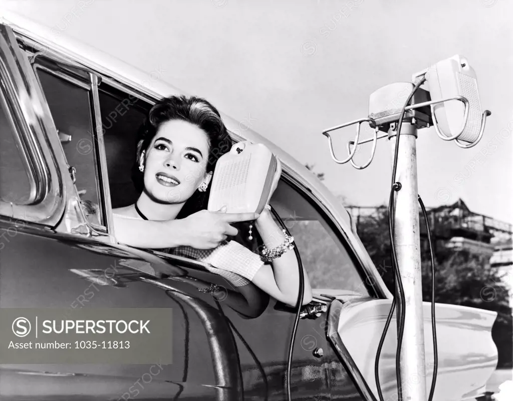 Hollywood, California: 1957. Natalie Wood displays the new drive in-movie speakers as she leans out of her car window.