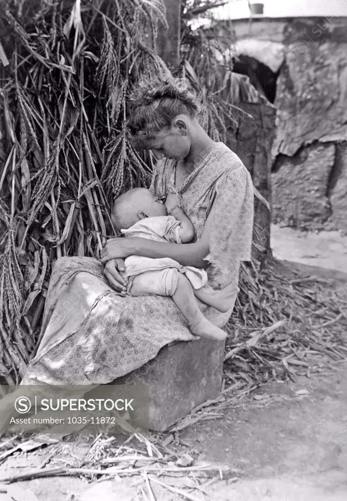 Hoopeston, Illinois:   1916. A mother sitting on a box next to cornstocks, breast feeding an infant.