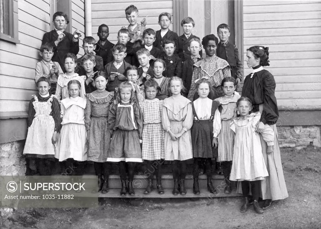 United States:  c. 1880. A racially mixed group of childen with their teacher pose for a portrait outside their schoolhouse. Many of them are holding their favorite items.