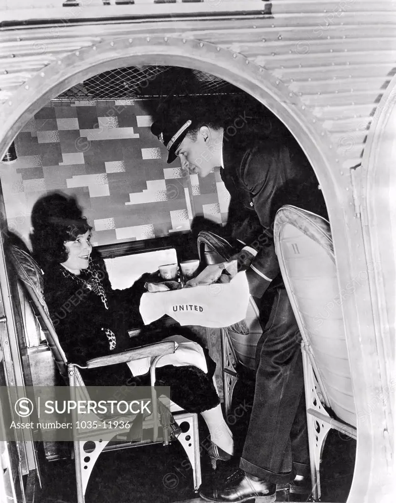 United States:  c. 1928. Interior view of a steward serving a passenger on a United Airlines plane.