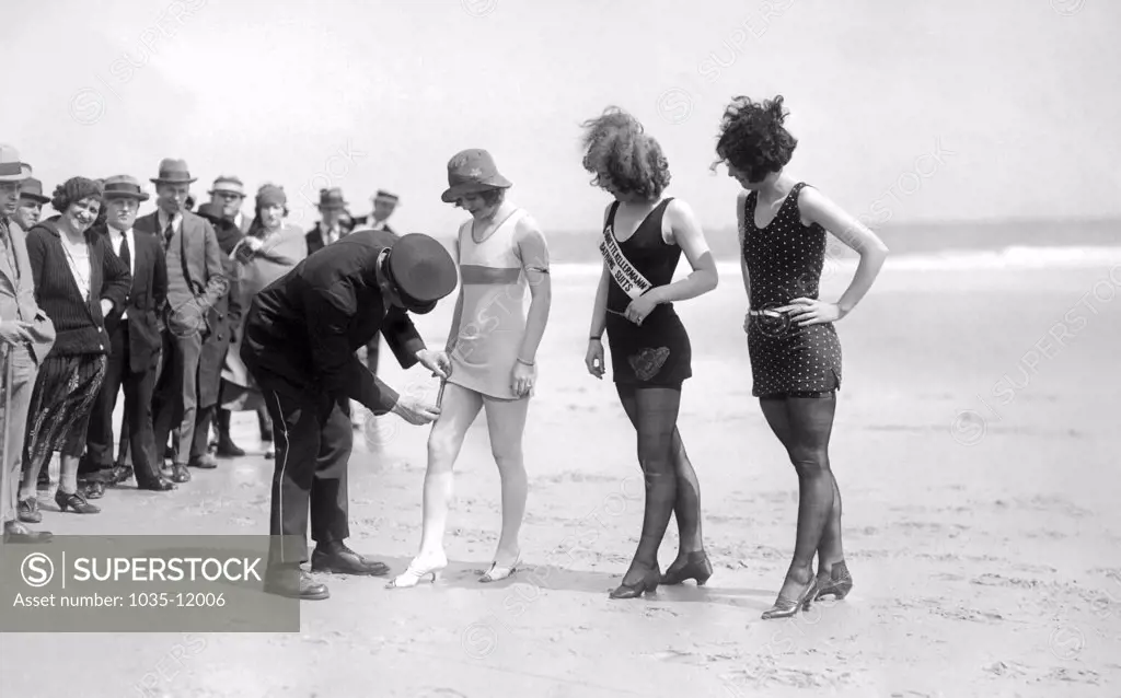 Atlantic City, New Jersey:  c 1921. A dubious and curious police officer measures the hem of the newest in bathing fashions. Are the suits decent  Time will tell!