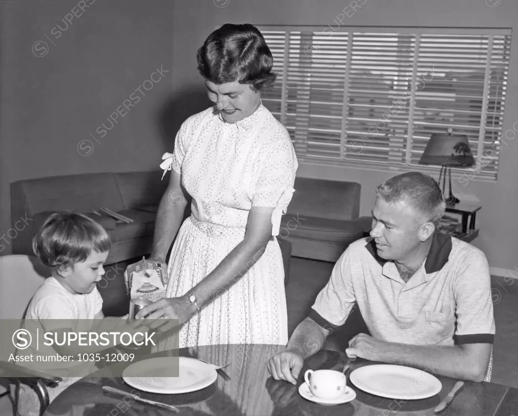 United States:  c. 1955. A mother pours a glass of milk for a young child at the kitchen table while the father looks on.