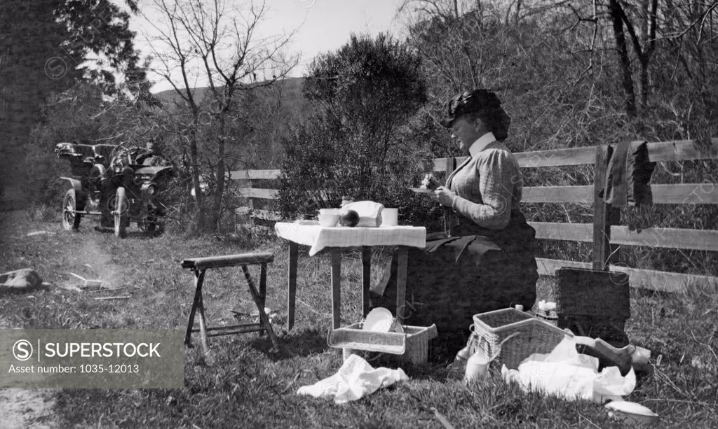 Portola Valley, California:   May, 1908. A woman in Victorian dress sits at a portable picnic table and eats lunch.