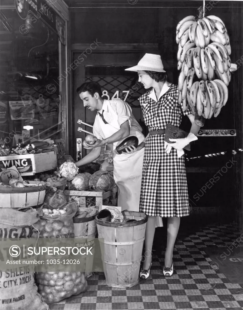 United States:  c. 1938. A stylish woman in a grocery store holds an eggplant while a clerk picks out a head of cabbage for her.
