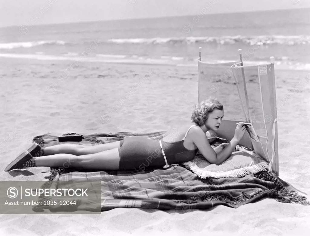United States: c. 1932. A woman lies comfortably on a beach with a large blanket, pillow, magazine holder, and wind screen.