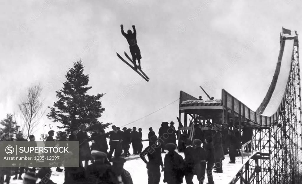 United States:  c. 1922. A photo showing a skier as he takes flight from a very tall and very narrow ski jump.