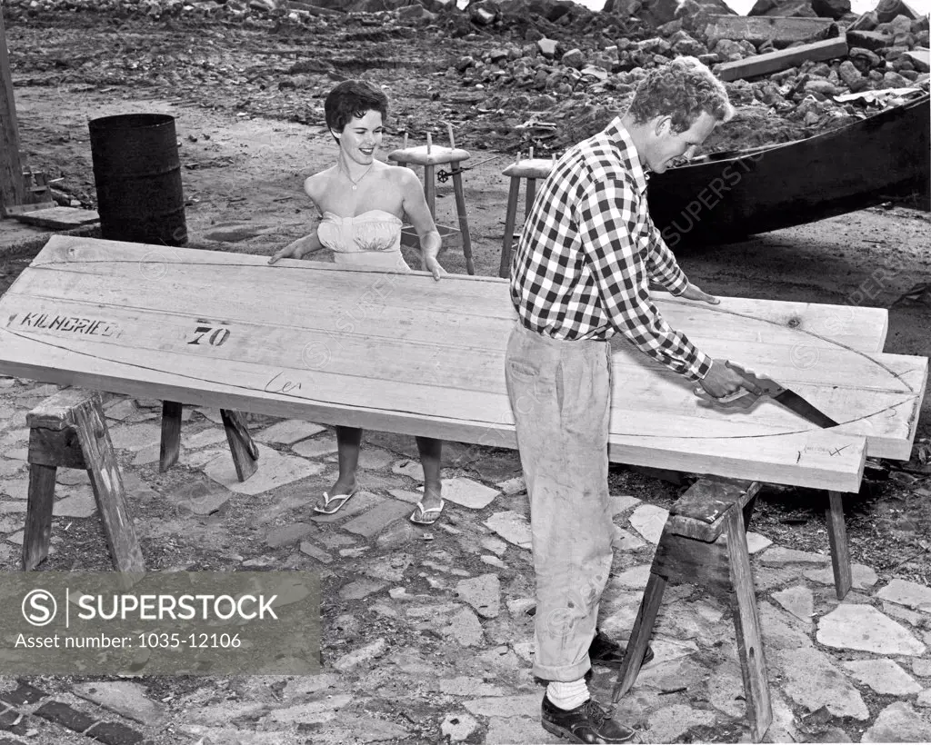 Santa Cruz, California:  c. 1959. A surfer cuts out his new board with a saw while his girlfriend holds things steady.