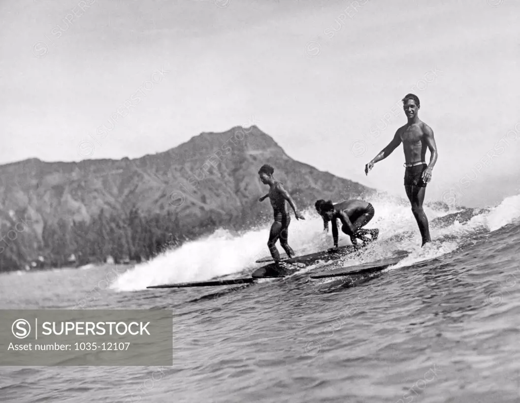 Honolulu, Hawaii:  c. 1926. Three native surfers ride their boards with ease at Waikiki Beach, with Diamond Head in the background.