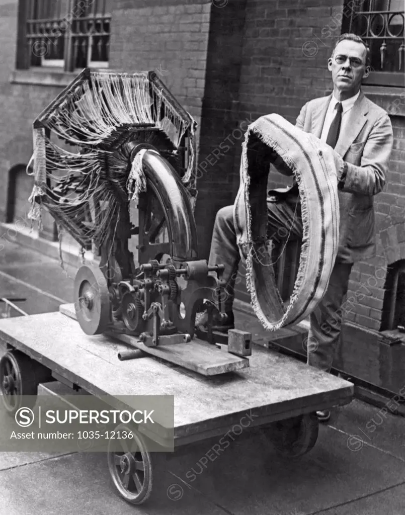 Washington, D.C.:  August 21, 1925. A Smithsonian Institute official holding the portable tire making device that was presented to them today. He's standing with the fabric weaving loom machine, and the second machine, which is not shown, constructs a three ply rubberized fabric. This machine makes it possible to set up small auto tire factories anywhere that's desired.