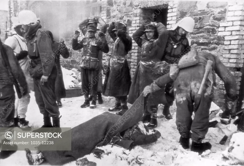 Germany:   February 14, 1945. An American soldier lifts the blanket from a fellow soldier to see if he was a friend, while captured German soldiers stand with their hands on their heads in this scene from somewhere on the U.S. First Army front.