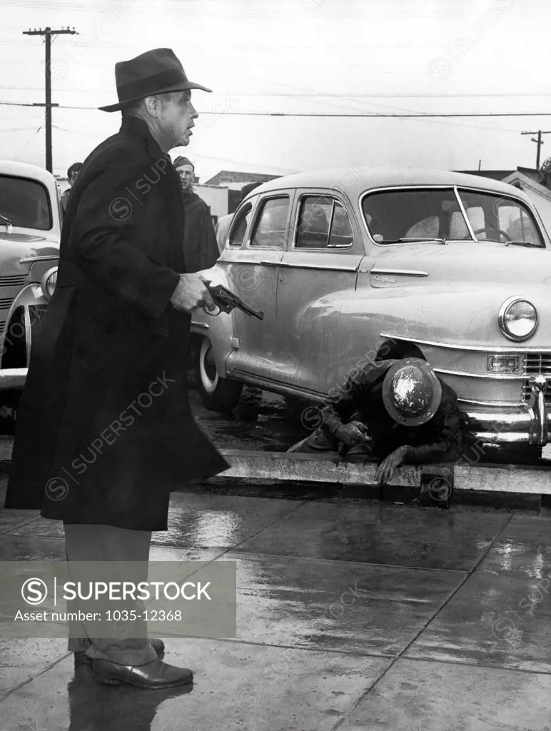 Los Angeles, California: October 1, 1946. A deputy sheriff stands with a cocked pistol near a fallen fellow officer, holding back a crowd of pickets demonstrating at the MGM Studio.