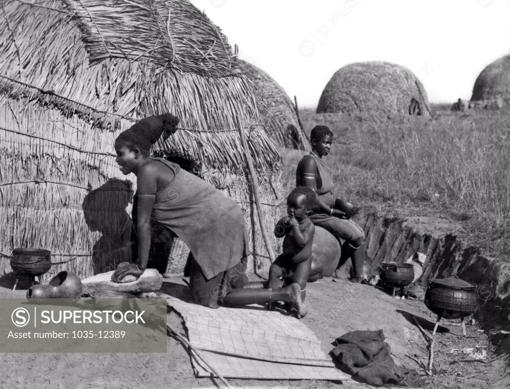 South Africa:  c. 1923. A native woman kneading bread on a flat stone outside her hut.