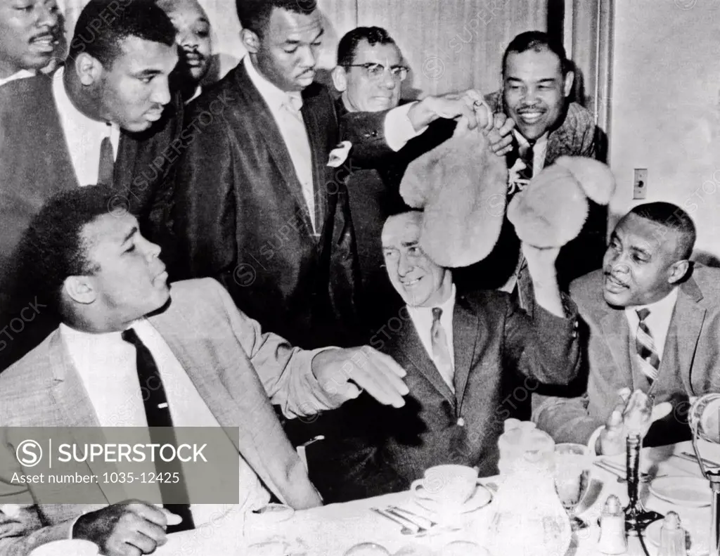 Denver, Colorado:  November 5, 1963. Cassius Clay recoils in mock horror from a pair of mink gloves that Champion Sonny Liston says he will use in their fight so that he won't hurt Clay's face. Joe Louis watches from the right rear.