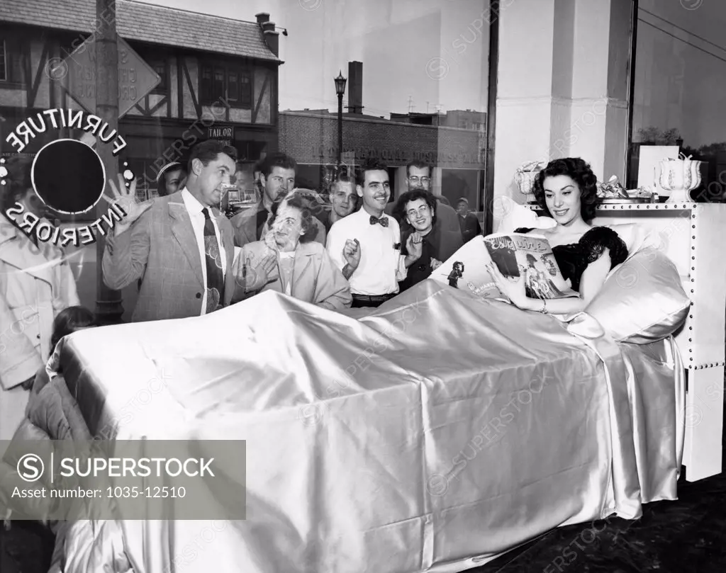 Chicago, Illinois:  October 10, 1951 A model reads an Esquire magazine in bed in the store window of Kay's Interiors to call attention to a new line of linens introduced by the store. She seems unperturbed by the rapt audience outside her window.