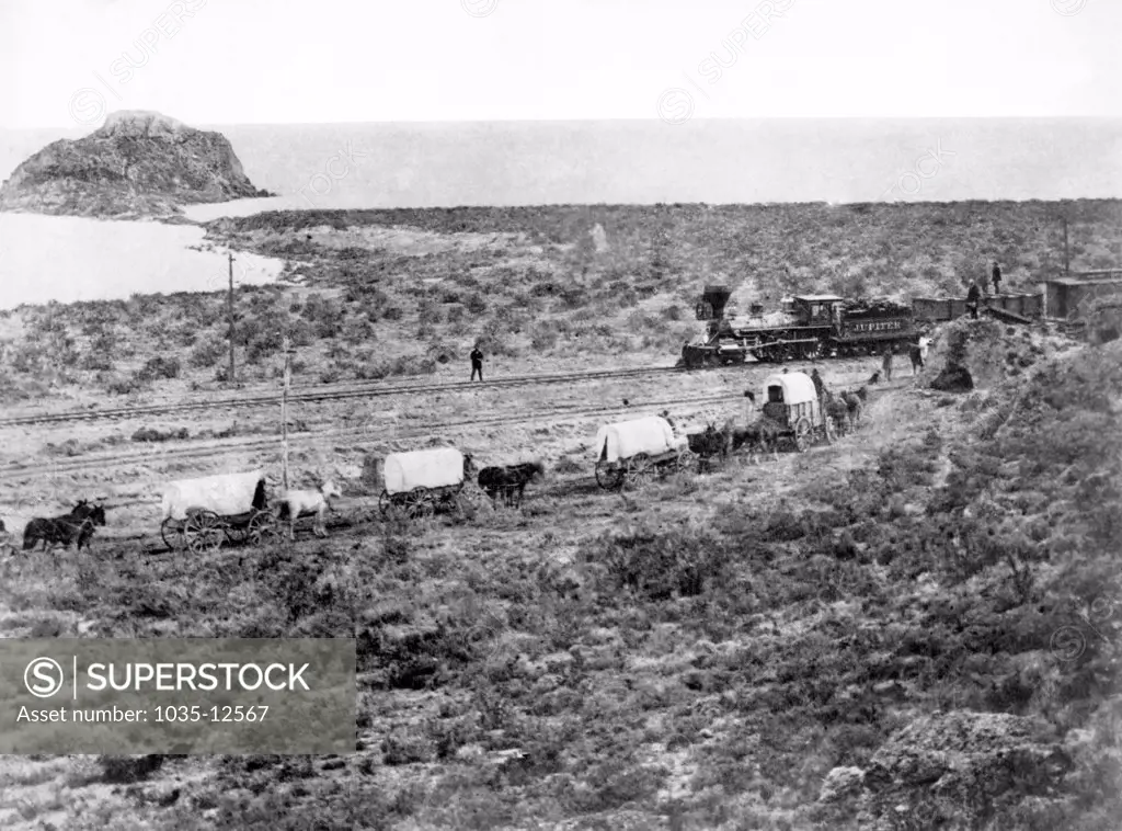 Monument Point, Utah:  c. 1868 The Central Pacific Railroad's Jupiter engine passes by a wagon train during the construction of the transcontinental railroad on the north side of the Great Salt Lake.