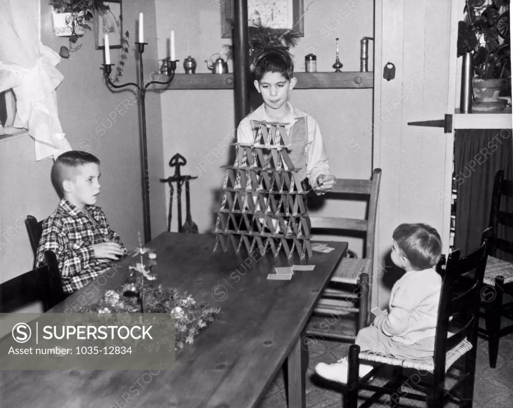 United States:  c. 1950. Two young siblings watch as their older brother puts the finishing touches on a towering pyramid of cards.
