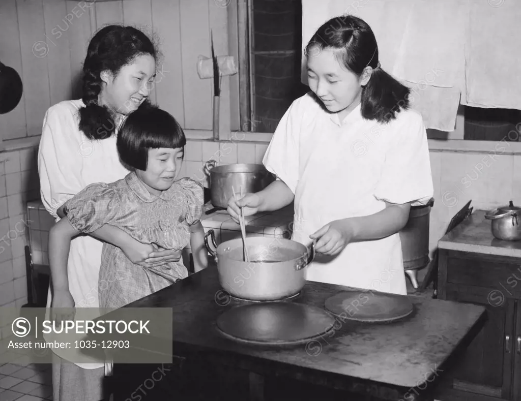 Nasu, Japan:  August 13, 1946 The three youngest daughters of Emperor Hirohito, on vacation at the Imperial Villa in the Japanese Alps, cook the evening meal for the seven household attendants as part of thier training in the domestic and culinary fields. Kazuko, 18, prepares the evening meal as Atsuko, 16, helps her younger sister, Takako, 8, get a peek at what is cooking.
