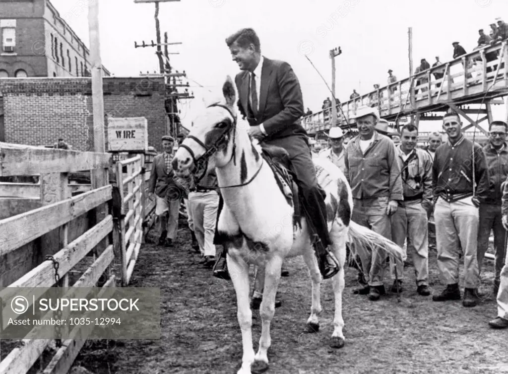 Sioux City, Iowa:  September 22, 1960 Workers smile and watch as Senator John F. Kennedy rides a white mule during a campaign visit to the stockyards here. He is on his way to Sioux Falls, South Dakota, to the National Plowing Matches.