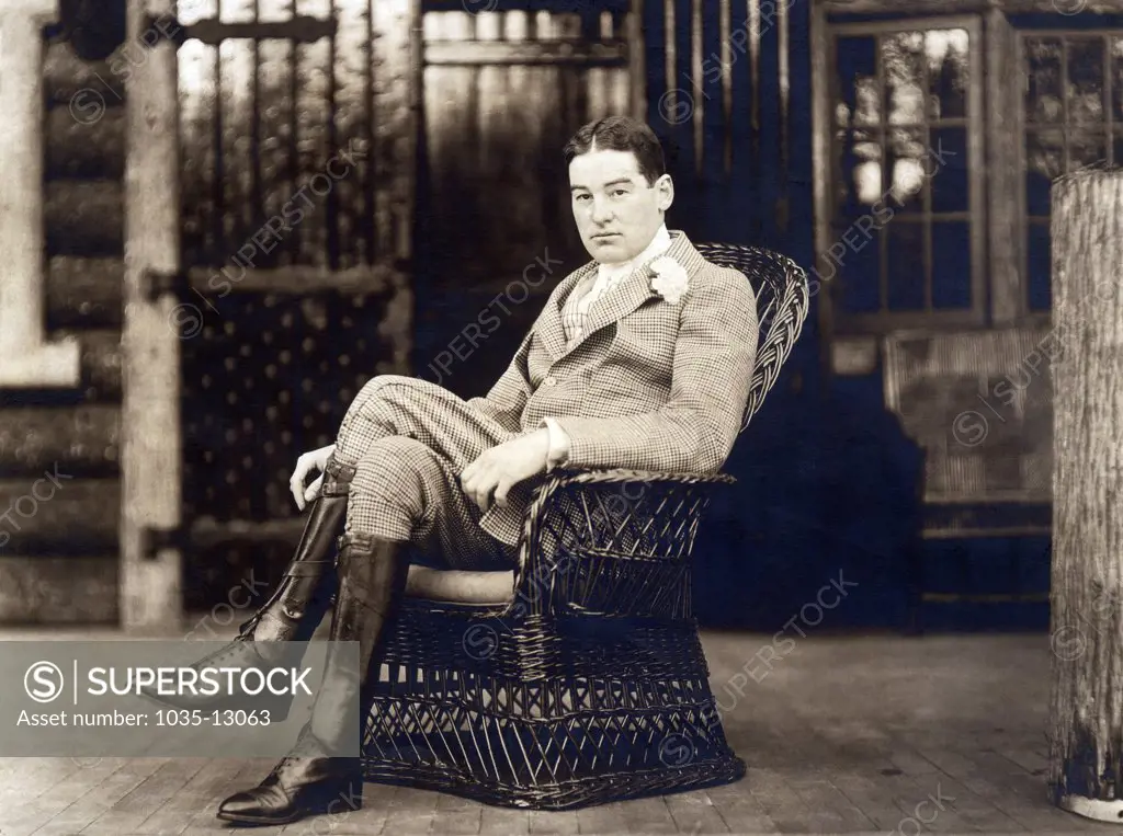 United States:  c. 1887 William K. Vanderbilt sits in a wicker chair wearing riding boots and carnation in his lapel.