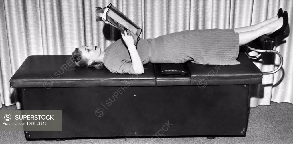 New York, New York:  April 20, 1955 A customer lays down on a reducing exercise table with a oscillating pad that lets her read as she loses weight. The Slenderella technique lets the pad under her hips do all the work.