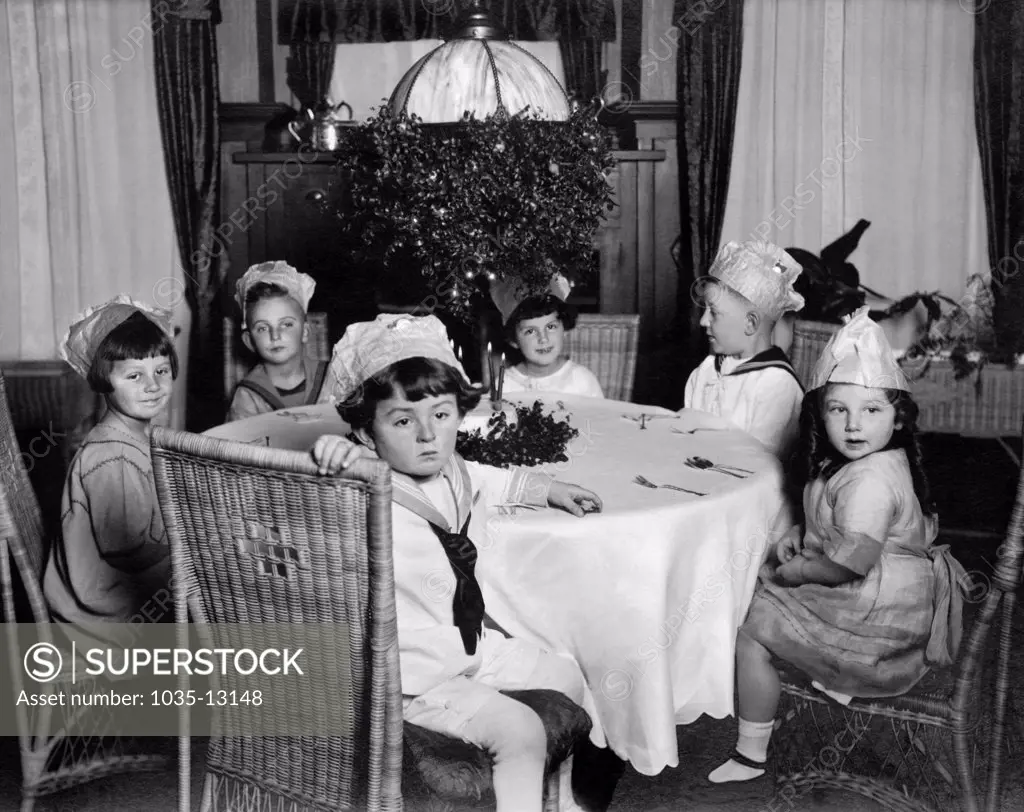 United States:  c. 1910. Six young children wearing party hats sitting around a table anxiously awaiting...