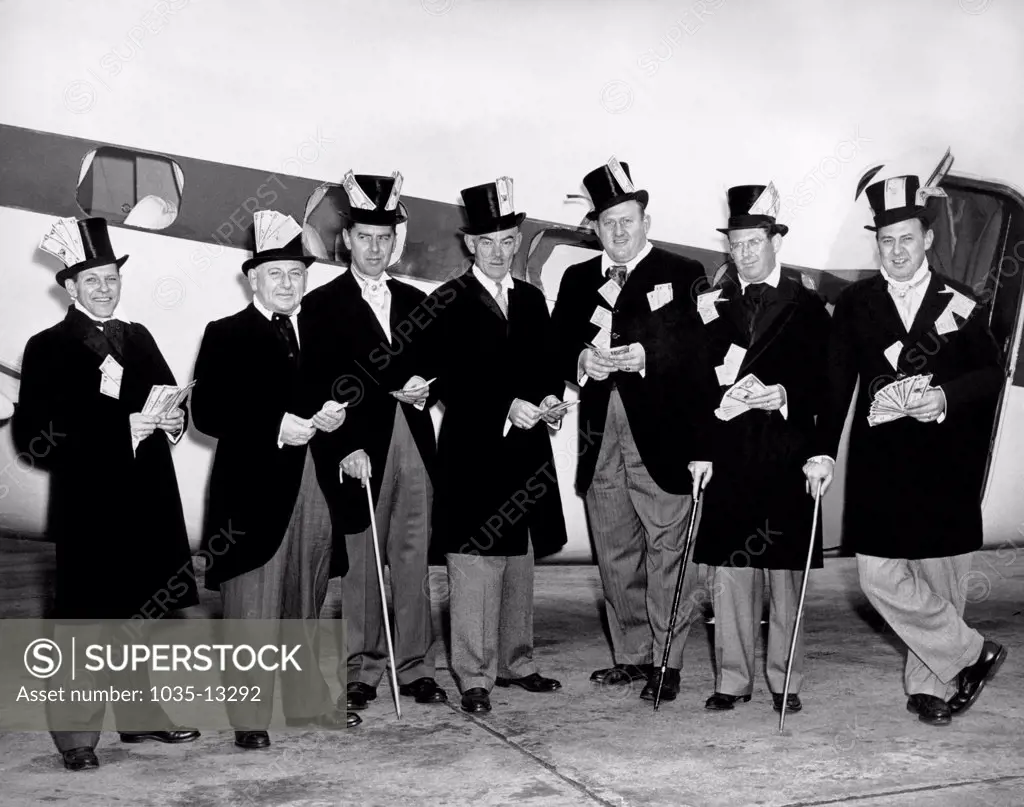 United States:  c. 1954. Seven men in tuxedos and top hats with handfuls of dollar bills in their hats and clothing pose next to an airplane.