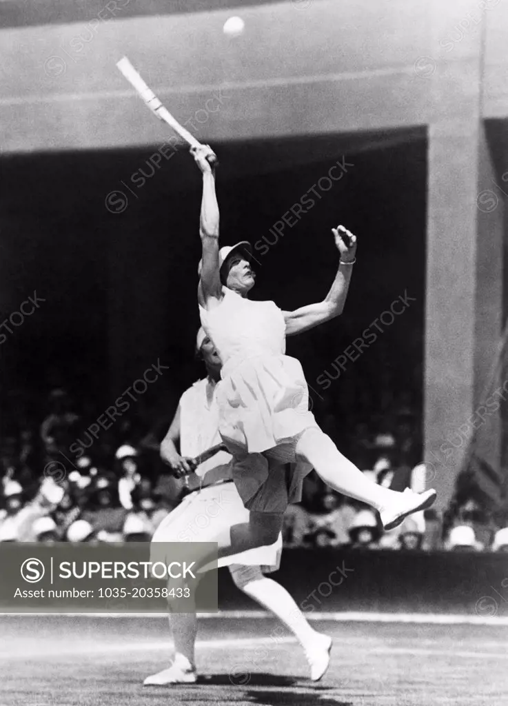 London, England:  1928 Kea Bouman of the Netherlands leaps high to try to return a high ball in the Doubles quarterfinals at Wimbledon. Her partner is British player Evelyn Colyer.