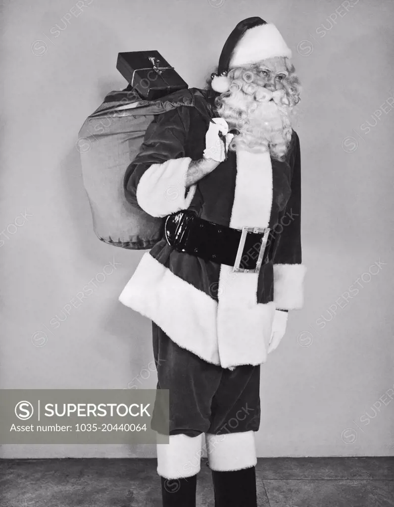 United States:  c.  1950 Santa Claus carrying a bag full of presents.