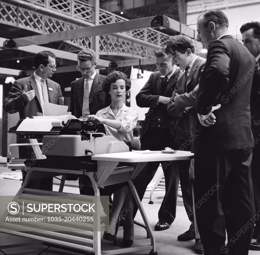 London, England:  c.  1962 A woman at Earl's Court Exhibition Hall demonstrates a new piece of office equipment while several men intently watch her.