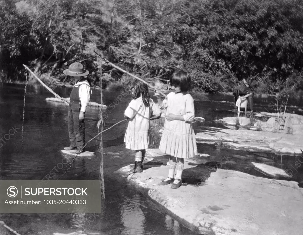 United States:   c. 1910 Young chidren try their luck at fishing in a small stream.