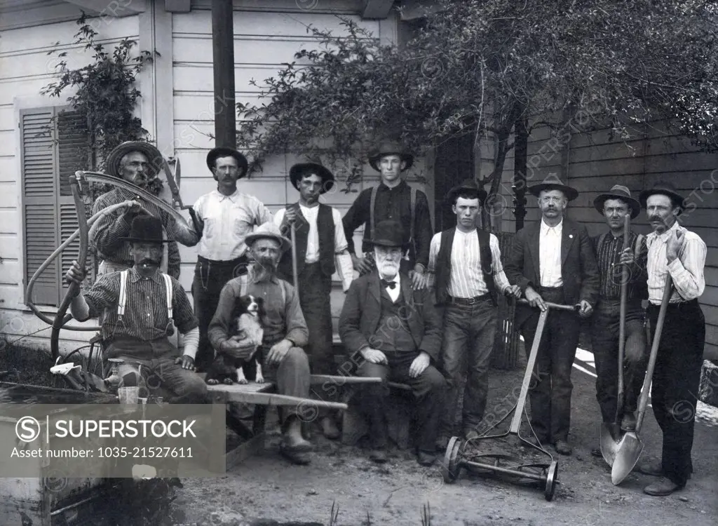 United States:  c. 1900 An older man with his workers and landscaping crew and their tools, including a wheelbarrow, shovels, shears, lawn mower and a scythe.