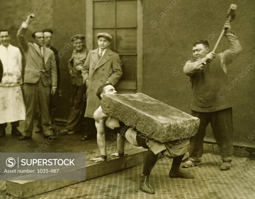Mid adult man raising a hammer to smash a slab of stone over a young man's chest