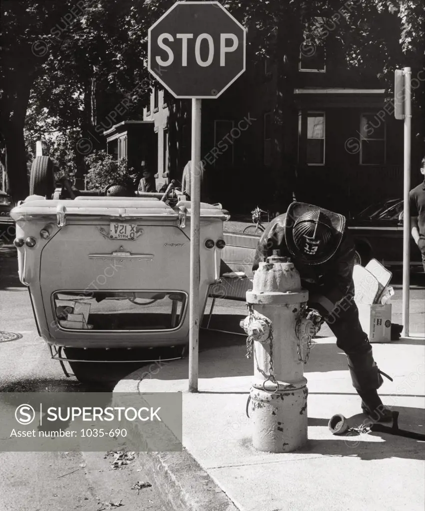 Firefighter opening a fire hydrant near an upside down car, 1967