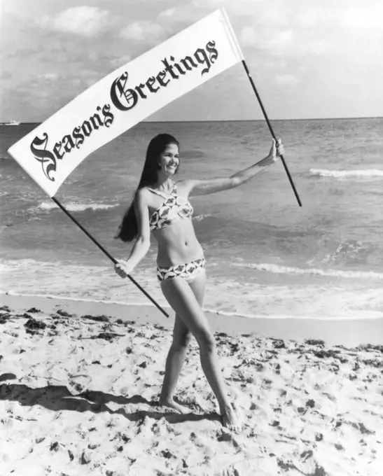 Miami Beach, Florida:  December 22, 1970 A young woman walking along the beach spreads holiday cheer with a banner and a brief bikini.