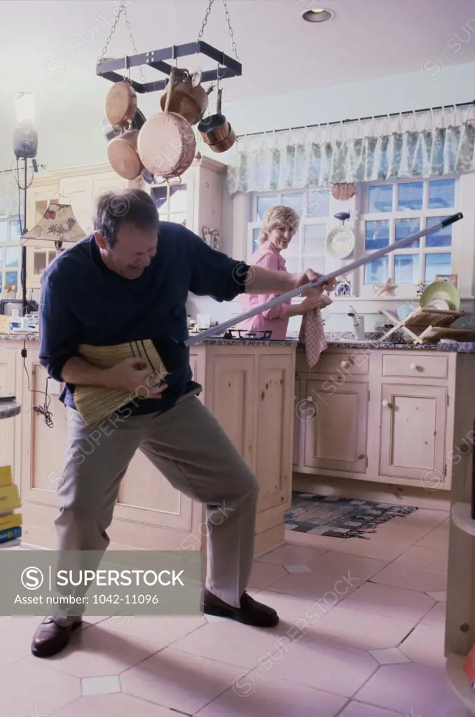 Mid adult man pretending to play the guitar with a broom and a mid adult woman standing behind him