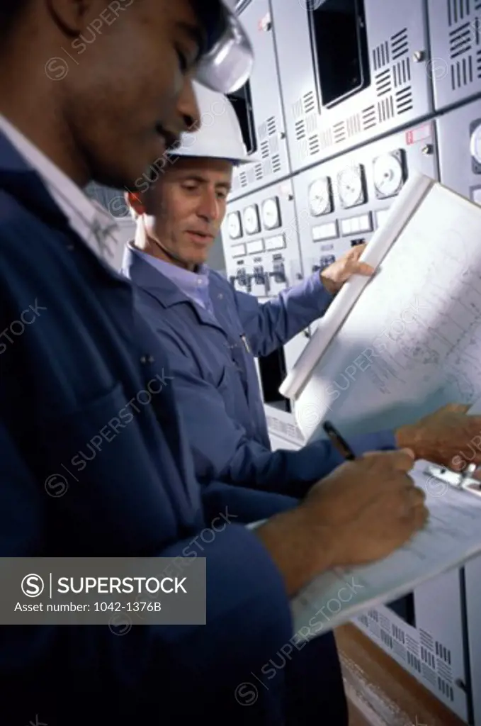 Side profile of two mid adult men standing in a factory holding blueprints