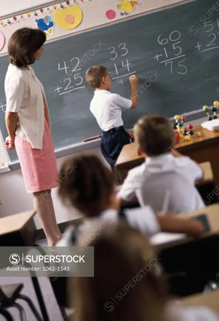 Boy writing on a blackboard in a classroom and a teacher standing beside him