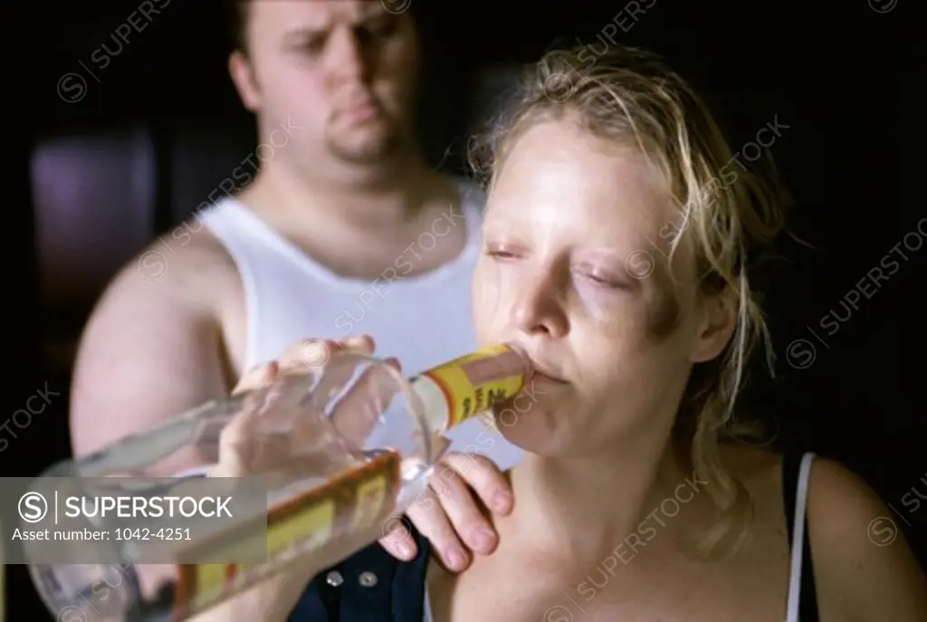 Young woman drinking alcohol from a bottle with a young man standing behind her