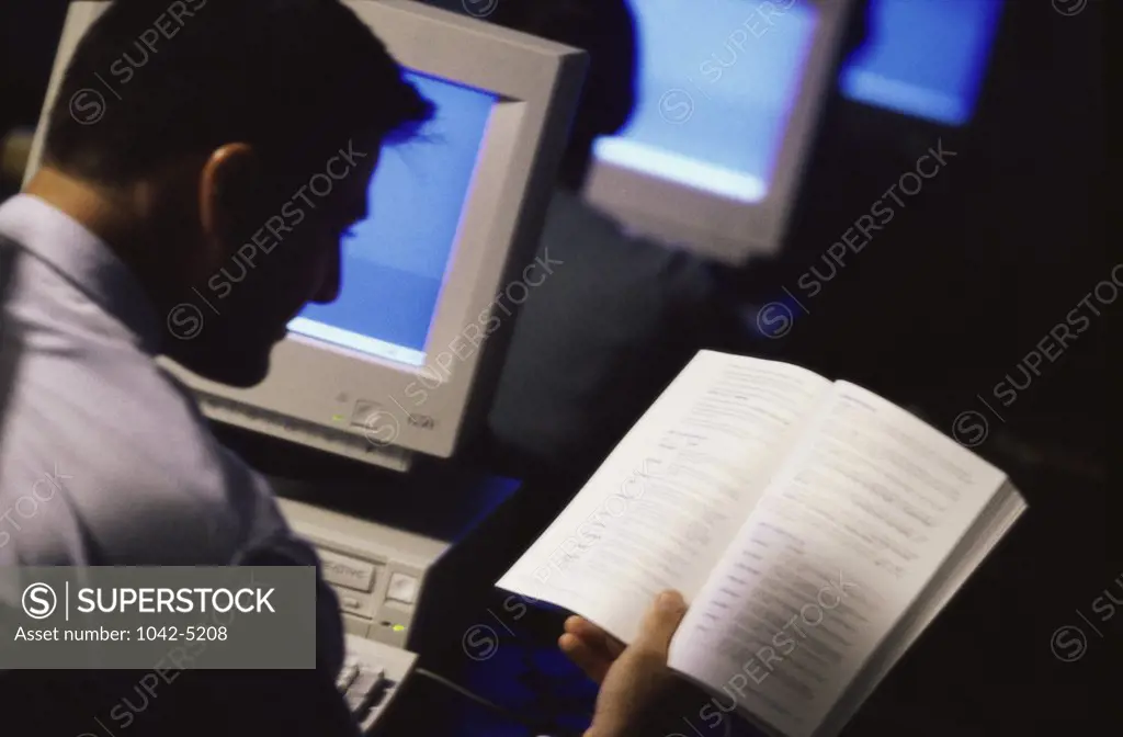 Rear view of a young man reading a book in front of a computer monitor