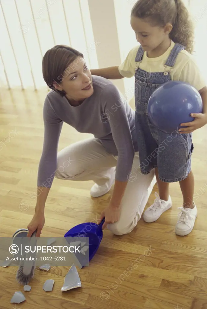 Mother sweeping up pieces of a broken vase with her daughter standing beside her