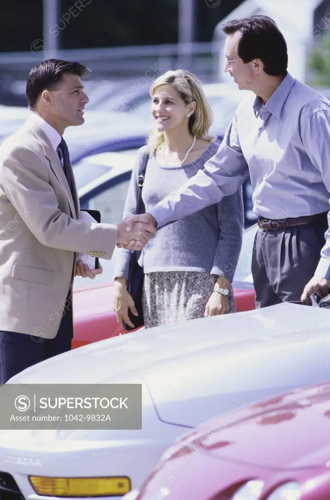 Two men shaking hands over the bonnet of a car with a woman standing besides them