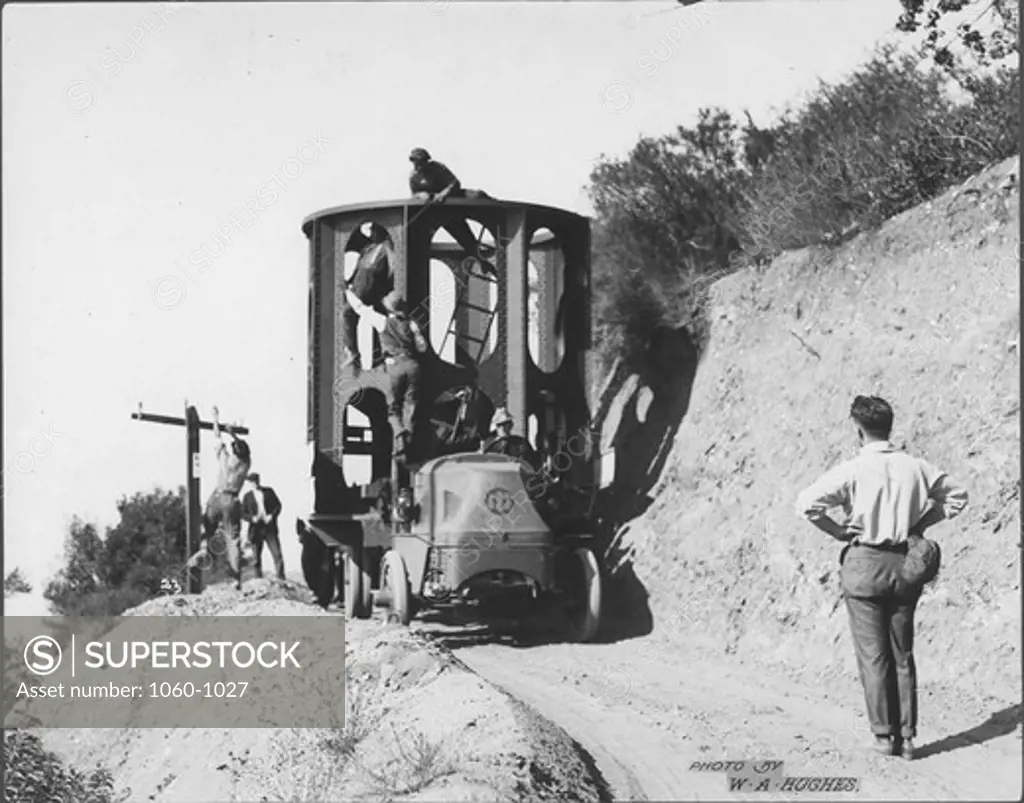 LOWER SECTION OF 100-INCH TELESCOPE TUBE BEING TRUCKED UP MT. WILSON TOLL ROAD.  MEN MOVING TELEPHONE WIRES OUT OF THE WAY.